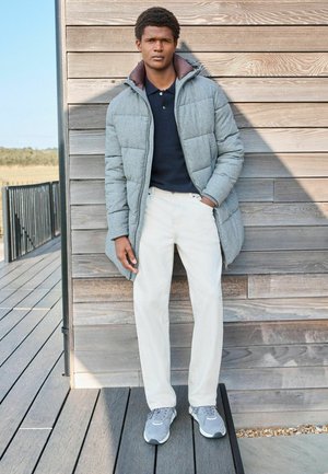 Chaqueta acolchada gris con un cuello marrón, camiseta polo azul marino, pantalones blancos y zapatillas grises, de pie contra una pared de madera.