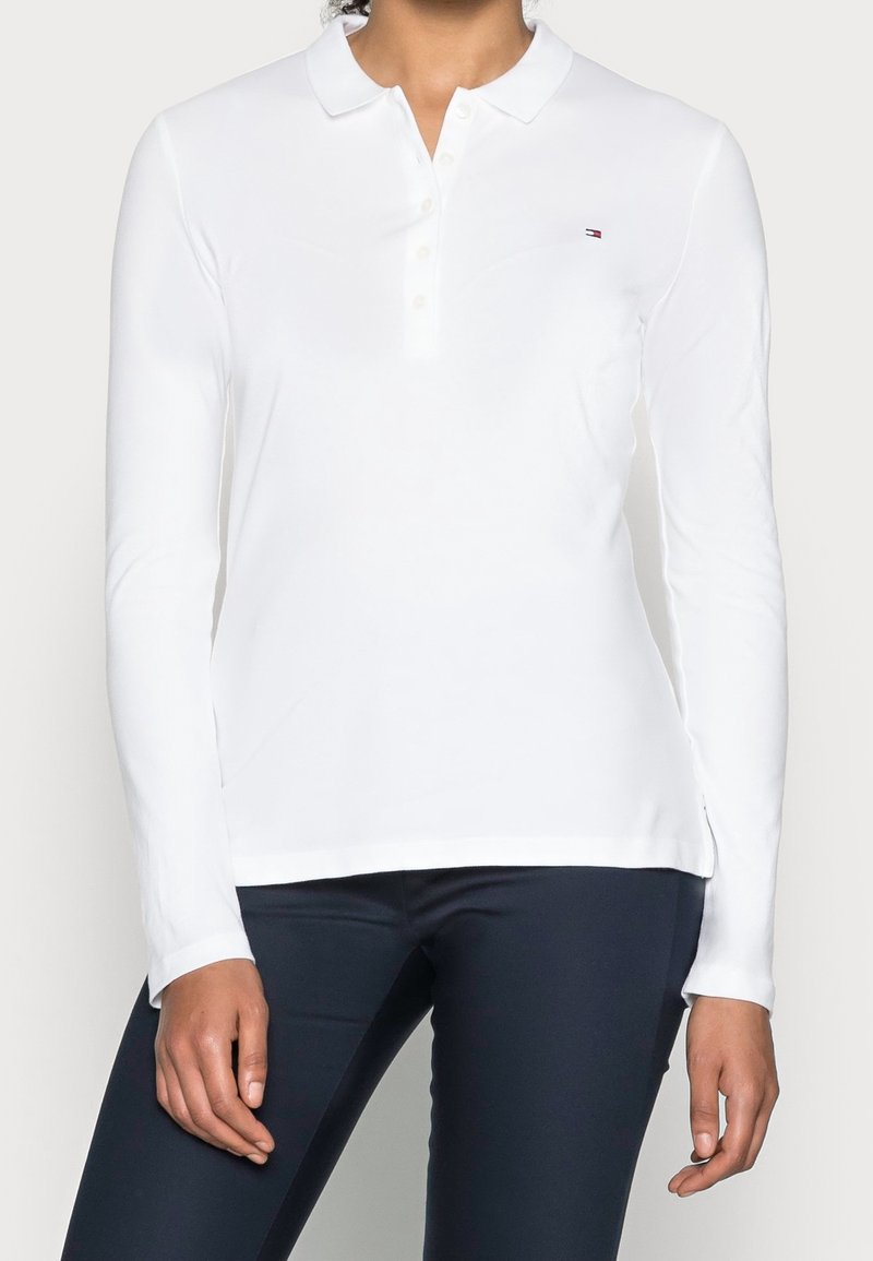 Femme portant un polo blanc à manches longues avec des boutons et un petit logo sur la poitrine, associé à un pantalon foncé.