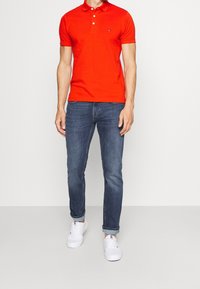 Camisa polo roja de manga corta con cuello clásico y tres botones, combinada con jeans oscurecidos y zapatillas blancas.