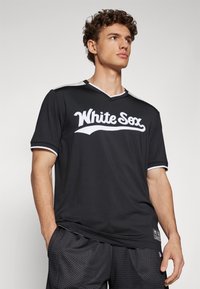 Crna sportska majica s bijelim natpisom "White Sox", s V izrezom, kratkim rukavima i bijelim detaljima na ramenima; nosi se s crnim teksturiranim šortsevima.