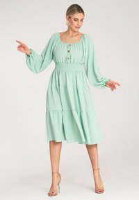 Miętowa sukienka z kwadratowym dekoltem, marszczonym pasem w talii, długimi rękawami i falbanistą spódnicą, z guzikami na przodzie.