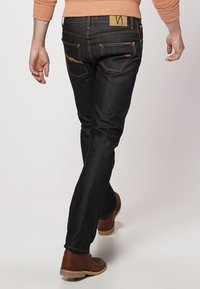 Mörk denimjeans med orange sömmar, slim fit. Bakvyn visar två fickor med böjda detaljer och ett tan-färgat loggopatch på midjebandet.
