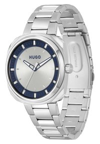 Reloj de acero inoxidable con una esfera en plata y azul marino, cristal mineral y pulsera de eslabones metálicos. Presenta la marca HUGO y marcadores de horas minimalistas.