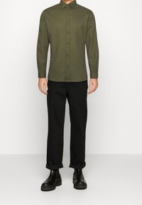 Oliwkowa zielona koszula z długim rękawem i kołnierzykiem na guziki, zestawiona z podwiniętymi czarnymi spodniami i masywnymi czarnymi botkami. Gładka tekstura tkaniny.