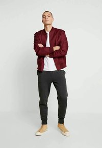 Tânăr stând cu brațele încrucișate, purtând o jachetă vișinie, un tricou alb, pantaloni negri și adidași bej, pe un fundal simplu.