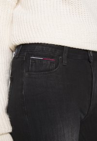 Jeans de mezclilla negros con un diseño ajustado, que cuentan con un único bolsillo delantero con un pespunte en rojo, blanco y azul. Combinados con un suéter de punto crema.