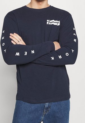 Mann trägt ein marineblaues Langarmshirt mit "Tommy"-Logo auf der Brust und "New York"-Schriftzug auf den Ärmeln, kombiniert mit blauen Jeans, die Arme verschränkt.