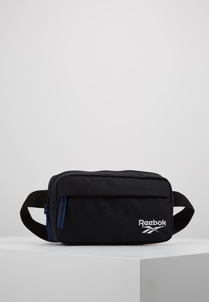 Bolsa de cintura negra Reebok con compartimentos con cremallera y correa ajustable, colocada sobre una superficie blanca frente a un fondo gris liso.
