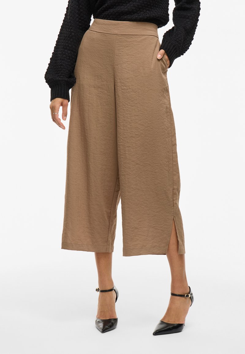 Bruna, vida byxor med en texturerad tyg, hög midja, sidofickor och slitsar i nederkant, matchade med svarta pumps med spetsig tå.