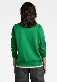 Sudadera de algodón verde con cuello redondo, mangas raglán y puños acanalados. Presenta una textura suave y un branding mínimo en el dobladillo.