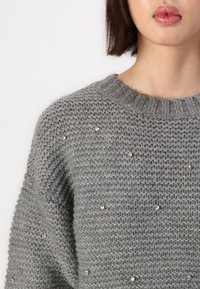 Donna che indossa un maglione grigio lavorato a maglia con piccoli strass decorativi, mostrando labbra, collo e spalla su sfondo bianco.