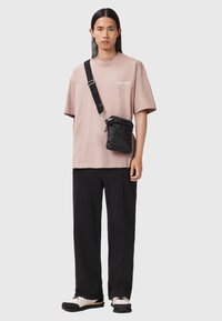 Beige T-Shirt mit kurzen Ärmeln und Logo, schwarze Umhängetasche mit gestepptem Muster, schwarze lässige Hose und schwarz-weiße Sneakers.