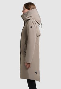 Beige padded coat with a high collar, adjustable drawstring hood, zippered pockets, and logo accents on the sides and chest.