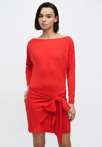 Czerwona sukienka z długim rękawem, dekoltem odkrywającym ramiona, o kroju kopertowym i zawiązaną kokardą w talii. Gładka tekstura tkaniny.