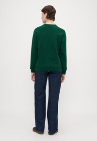 Grønn hettegenser laget av myk stoff, med rund hals og ribbestrikkede ermer. Kombinert med mørkeblå rette jeans.