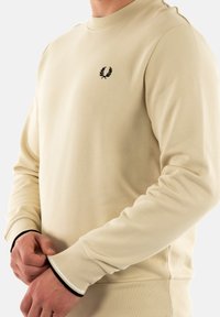 Sudadera beige con cuello redondo y puños acanalados elegantes. Presenta un logo negro bordado en el pecho. Textura suave.