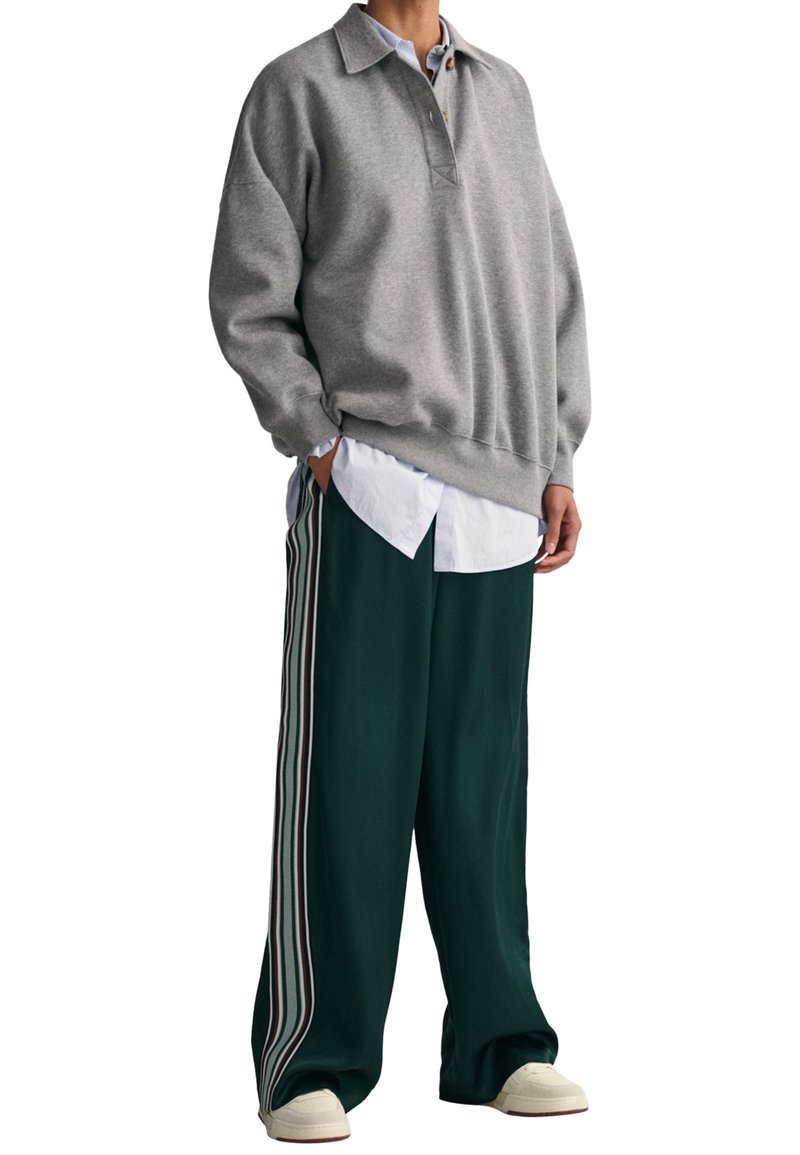 Grauer Pullover mit Kragen, getragen über einem weißen Hemd. Grüne Hose mit drei vertikalen Streifen auf jeder Seite. Weiße Schuhe runden den Look ab.