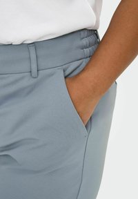 Lys blå bukser med elastisk midjebånd, sidelommer og glatt stofftekstur. Nærbilde av lommen og midjeområdet.