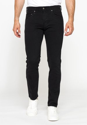 Uomo con jeans neri slim fit e sneakers bianche in piedi davanti a uno sfondo chiaro e uniforme, mani rilassate lungo i fianchi.