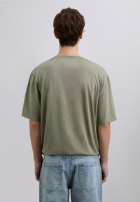Camiseta de algodón verde oliva con un ajuste relajado, mangas cortas y escote redondo. La vista trasera muestra una textura lisa y detalles de costuras mínimas.