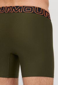 Olive green boxer shorts with a black waistband featuring "ARMOUR" in bold orange lettering. Smooth fabric with a fitted design.