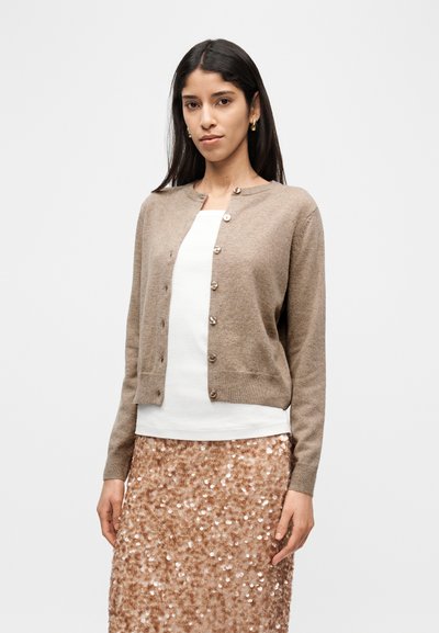 Χακί ζακέτα με στρογγυλή λαιμόκοψη και κουμπιά, σε συνδυασμό με λευκή μπλούζα και μια λαμπερή ροζ φούστα με παγιέτες.