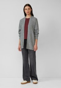 Grauer Strickcardigan mit offener Vorderseite, kombiniert mit einem kastanienbraunen Oberteil und schwarzen, weiten Hosen. Die Schuhe haben ein Design in Weiß und Braun.