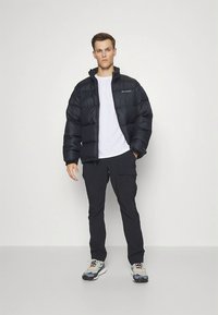 Černá péřová bunda s vysokým límcem, bílé tričko a černé kalhoty. Šedé a oranžové tenisky doplňují outfit.