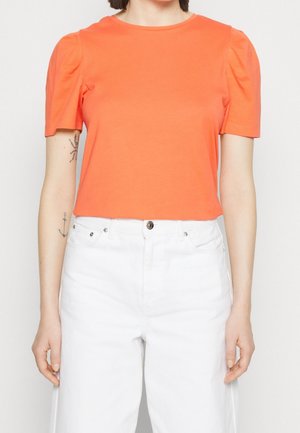 Vrouw draagt een feloranje top met korte mouwen en pofschouders en een witte broek met hoge taille, staand tegen een effen achtergrond.