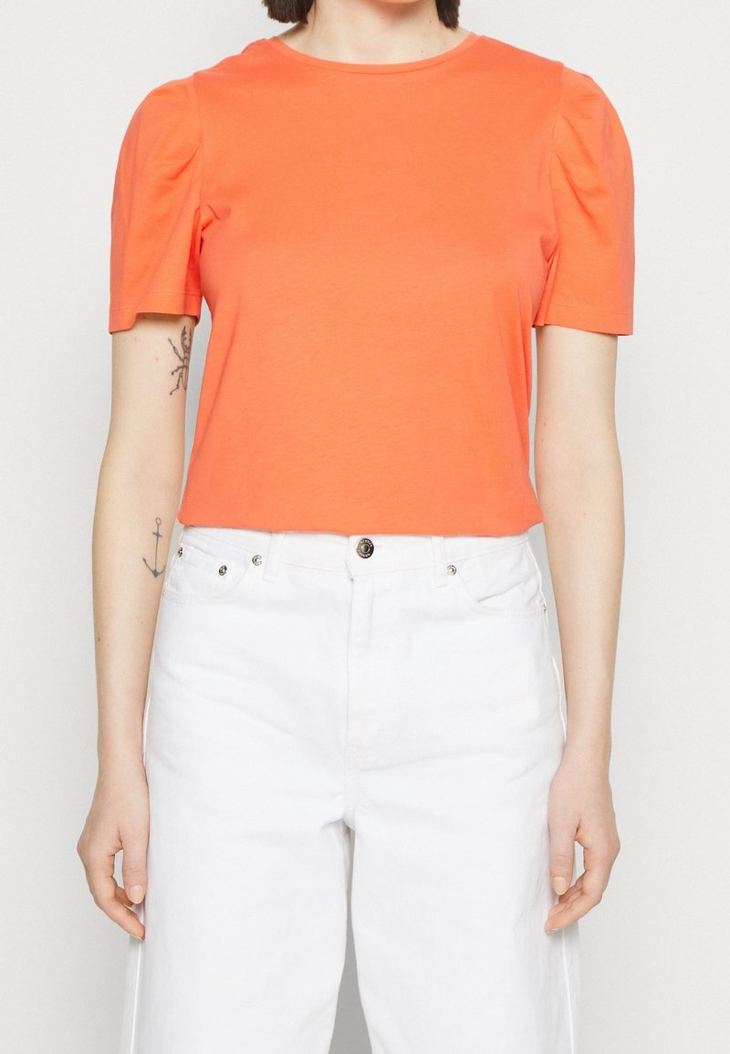 Femme portant un top court orange vif à épaules bouffantes et un pantalon blanc taille haute, debout devant un fond uni.
