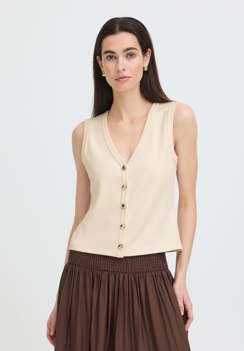 Femme aux longs cheveux foncés portant des boucles d'oreilles anneaux dorées, un haut beige sans manches avec des boutons, et une jupe marron taille haute sur un fond uni.