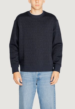 Felpa blu navy con un motivo logo texture, collo a giro, e polsini a costine. Abbinata a jeans denim blu chiaro.