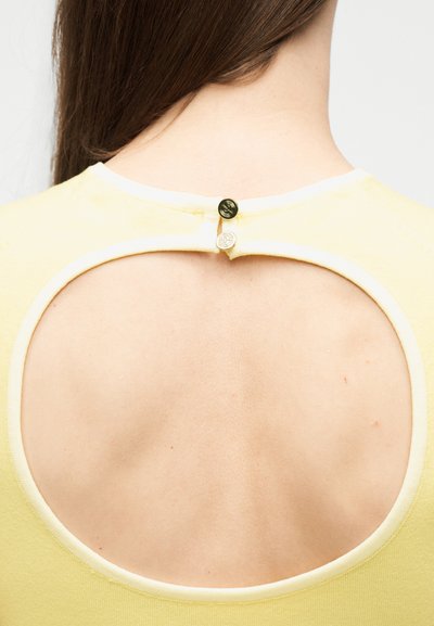 Πίσω όψη ενός ατόμου που φοράει κίτρινο τοπ με μεγάλο κυκλικό άνοιγμα και δύο μικρά κουμπιά που στερεώνουν το ντεκολτέ.