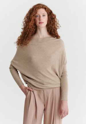 Mujer con cabello largo rizado y rojo, vistiendo un suéter beige holgado y pantalones anchos marrón claro sobre un fondo blanco liso.