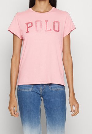 Femme portant un T-shirt à manches courtes rose clair avec "POLO" en grandes lettres roses délavées et un jean bleu dégradé.