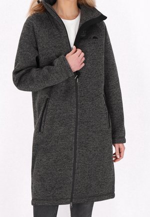 Vrouw draagt een donkergrijze fleecejas tot op de knie met een ritssluiting en twee zijzakken, houdt de rits bij de borst vast, witte blouse eronder.