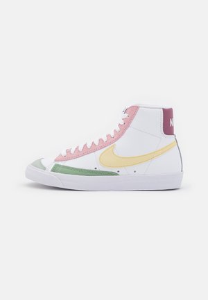 Wysokie buty sportowe Nike z białą podstawą, różową panelem z dziurkami, żółtym logo Swoosh, zielonym detalem na palcu i bordową zakładką na pięcie. Biała podeszwa i sznurówki.