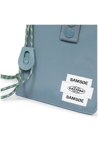 Borsa in tessuto blu con superficie testurizzata, caratterizzata da cordino intrecciato verde e grigio, chiusura con bottone a pressione e doppie etichette del marchio bianche.