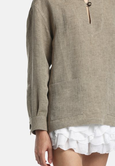 Laza bézs lenvászon hosszú ujjú felső elülső zsebekkel és gombos mandzsettával, fehér rétegzett fodros szoknya vagy rövidnadrág fölött viselve.