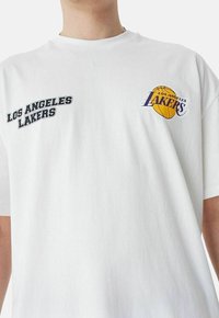 Camiseta blanca de algodón con "LOS ANGELES LAKERS" en texto negro a la izquierda y un logo a color a la derecha. Corte relajado y mangas cortas.