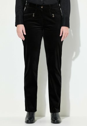 Persona che indossa pantaloni neri aderenti con tasche frontali con zip, camicia nera con bottoni e stivaletti neri, in piedi su sfondo bianco.