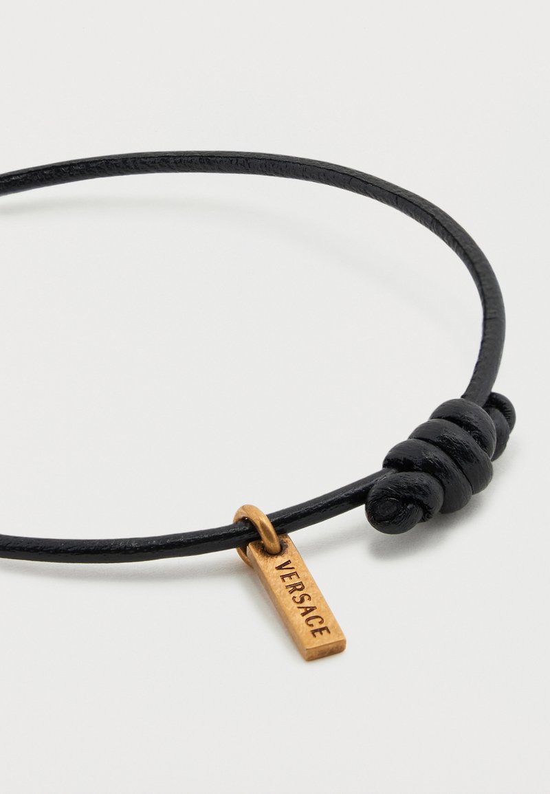 Black leather cord bracelet with adjustable knot and small gold rectangular charm engraved with "VERSACE" lettering.