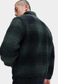 Zielona polarowa kurtka z wysokim kołnierzem, teksturowana powierzchnia oraz kratkowany wzór w różnych odcieniach zieleni. Posiada długie rękawy.