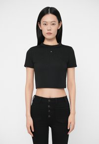 Γυναίκα με ίσια μαύρα μαλλιά, φορώντας μια μαύρη κομμένη τοπ με κοντούς μανίκους και ψηλόμεσο μαύρο παντελόνι με λεπτομέρειες κουμπιών.