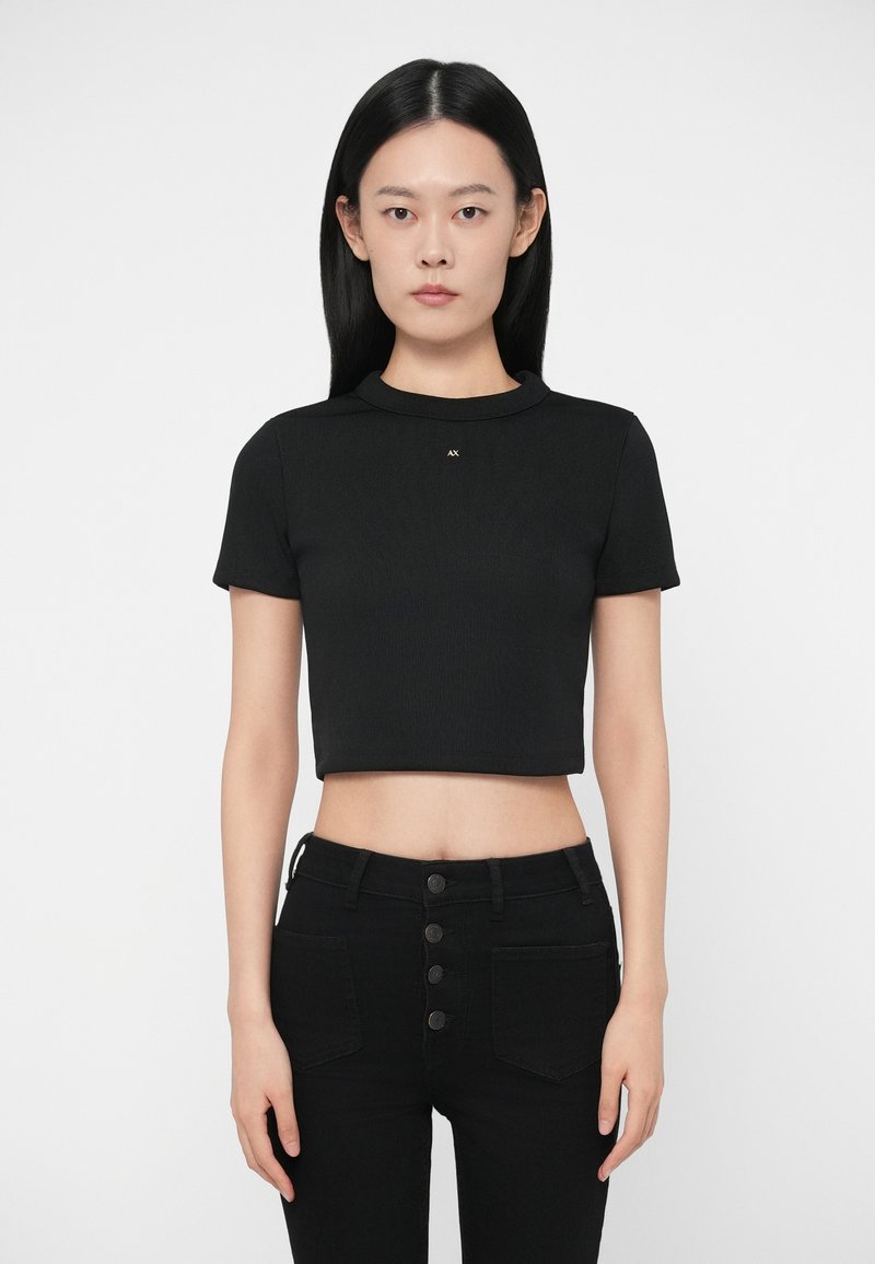 Γυναίκα με ίσια μαύρα μαλλιά, φορώντας μια μαύρη κομμένη τοπ με κοντούς μανίκους και ψηλόμεσο μαύρο παντελόνι με λεπτομέρειες κουμπιών.