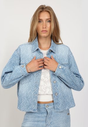 Blonde Frau trägt hellblaue gemusterte Jeansjacke und passende Hose über weißem gestricktem Croptop, richtet den Kragen der Jacke.