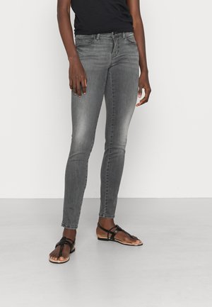 Jean gris slim porté avec des sandales plates noires à brides et un top noir, présenté de la taille aux pieds sur un fond uni.