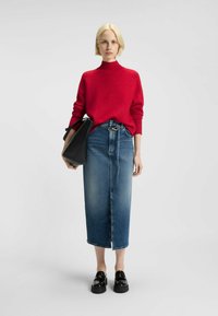 Pull en tricot rouge avec col montant, assorti d'une jupe midi en denim bleu avec une fente frontale. Le modèle tient un sac fourre-tout noir. Chaussures à plateforme noires.