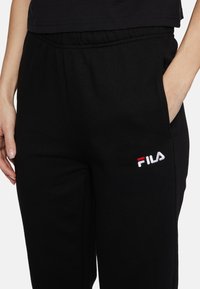 Zwarte trainingsbroek met elastische tailleband, zijzakken en rood-wit geborduurd Fila-logo op de dij. Soepele stoftextuur.