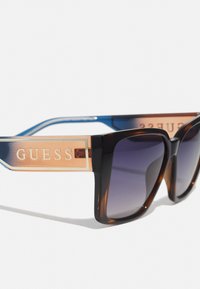 Occhiali da sole oversize marroni e neri con lenti sfumate e aste decorate con dettagli dorati che presentano la scritta "GUESS" in lettering in rilievo.